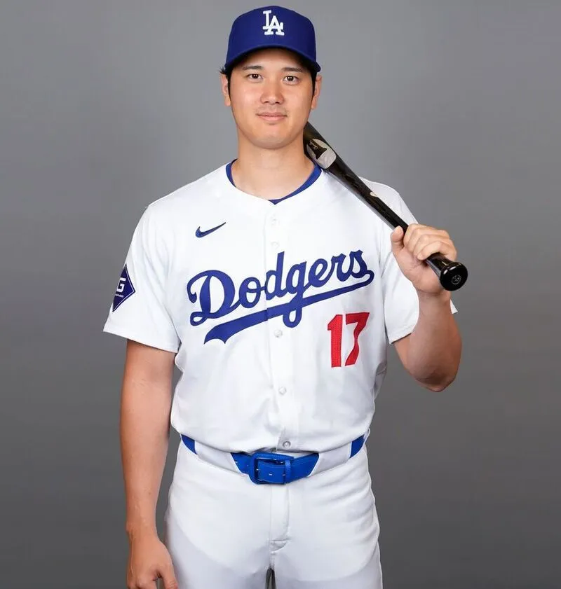 player wearing custom white baseball uniform blue printing on front holding bat in hand with grey background